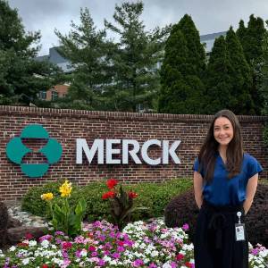 Lauren Favell Success Story Lauren Favell stands in front of a flower bed and a large sign that says Merck.